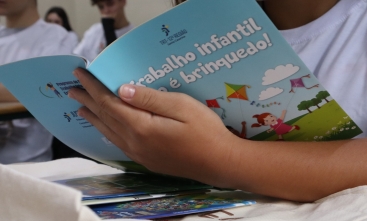 Imagem em close de um jovem segurando uma cartilha com o nome Trabalho infantil não é brinquedo