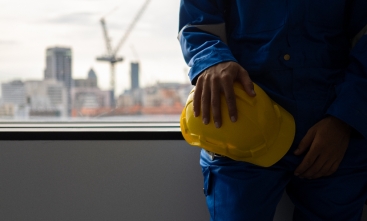 Trabalhador de obra com uniforme azul segura capacete amarelo diante de janela com cidade ao fundo