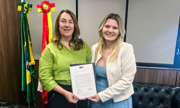 Duas mulheres vestidas formalmente e sorridentes seguram um documento dentro de uma sala