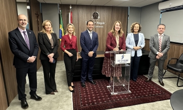 Três homens e quatro mulheres em pé, em uma sala, lado a lado, todos sorridentes. Uma das mulheres assina um documento em um púlpito transparente