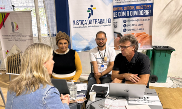 Foto de dois homens e uma mulher prestando atendimento a uma mulher loira. Atrás da mesa, um banner da Justiça do Trabalho