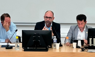 Homem calvo, de óculos, sentado à frente de um notebook, faz uma apresentação. Ao lado dele estão outros dois homens.