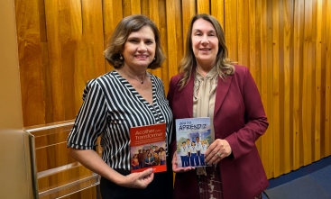 Duas mulheres posam para a foto. Cada uma exibe uma cartilha