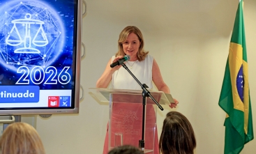 Mulher loira, vestindo blusa branca e saia rosa, fala ao microfone em uma sala de aula. Ela está levemente sorrindo.