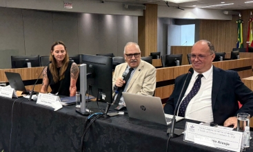 Três palestrantes sentados à mesa com microfones em auditório institucional