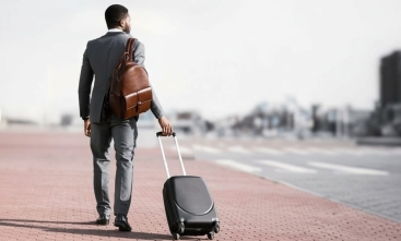 Fotografia ilustrativa de homem negro de terno claro e malas olhando para o horizonte distante
