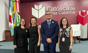 Três mulheres vestidas formalmente e um homem de terno azul marinho posam para a foto, sorridentes, em um auditório. Ao fundo, a logomarca da Universidade Católica