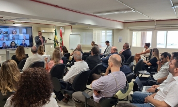 Homem dá palestra a público sentado em sala; bandeiras ao fundo e telão com videoconferência