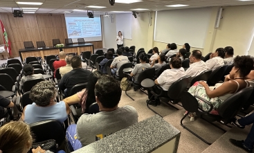 Foto de uma palestra, com pessoas sentadas em um auditório a uma mulher palestrante na frente do público 