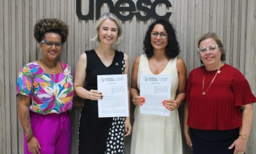 Quatro mulheres estão em pé, posando e sorrindo para a foto, segurando um contrato