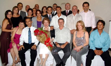 Foto posada de 21 pessoas, algumas sentadas e outras em pé. No centro, Ione Ramos está sentada e segurando, sorridente, uma buque de flores