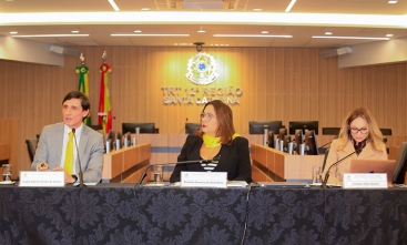 Um homem e duas mulheres estão sentados em uma mesa de honra, durante uma palestra