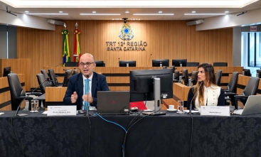Duas pessoas, um homem e uma mulher, sentados diante de mesa de honra durante palestra em plenário