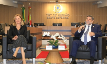 Home de terno escuro fala ao microfone sentado em uma poltrona, na sala de sessões do Pleno do TRT-SC. Ao lado dele, em outra poltrona, uma mulher loira sorridente, com roupa preta