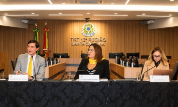 Um homem e duas mulheres estão sentados em uma mesa de honra, durante uma palestra