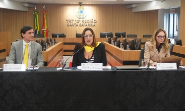 Um homem e duas mulheres estão sentados em uma mesa de honra, durante uma palestra