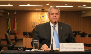 Fotografia de homem sentado falando em uma mesa de honra, durante uma palestra
