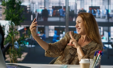 Fotografia ilustrativa de mulher sorrindo para o próprio celular em ambiente de escritório
