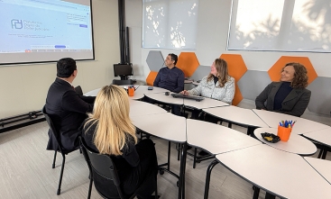 Grupo de três pessoas e dois homens sentados à mesa observando um telão. Uma das mulheres mexe no teclado e esboça um leve sorriso
