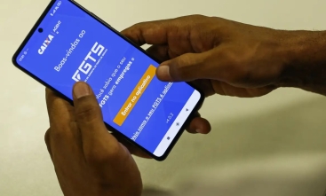 duas mãos segurando um celular, com o aplicativo da "caixa" aberto, tendo o fundo azul, com um letreiro em branco escrito "FGTS"
