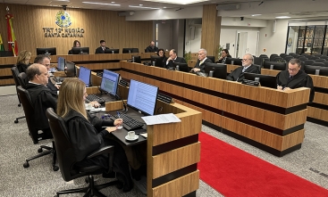 13 pessoas vestem roupas pretas, todas sentadas em torno de duas bancadas laterais e uma central, formando a letra U