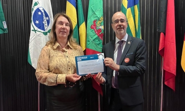 Uma mulher vestida formalmente e um homem de terno e gravata seguram, juntos, um certificado. Ao fundo bandeiras do Brasil e dos estados do Paraná, Santa Catarina e outra não identificada