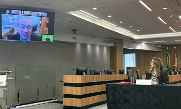 Mulher sentada à mesa de um evento observa palestrante que aparece em um telão