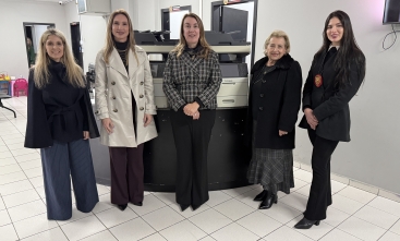 Cinco mulheres posam para a foto. Atrás delas, dois equipamentos de informática