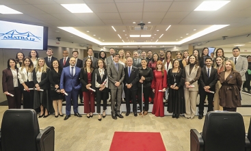 Foto de grupo com cerca de 40 pessoas, entre homens e mulheres, reunidas em um auditório. A maioria está em pé, lado a lado, em frente a uma tela com o logotipo da Amatra 12. As pessoas vestem roupas sociais e algumas seguram certificados. Ao fundo, há cadeiras e equipamentos de apresentação.