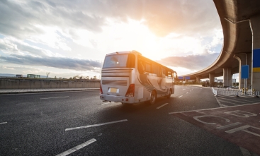onibus branco, em uma estrada, com o céu de fundo possuindo sol e nuvens