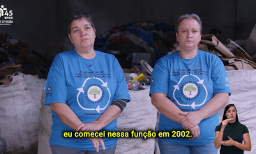 Duas mulheres brancas, uma de cabelos escuros e outra loira, estão lado a lado, olhando para a câmera, à frente de resíduos para reciclagem