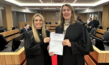Duas mulheres vestindo toga, numa sala de sessão de julgamentos, exibem um documento. Ambas estão sorridentes