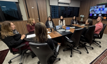 Grupo de sete mulheres e um homem, todos sentados ao redor de uma mesa. Eles observam um telão onde aparecem pessoas em videoconferência