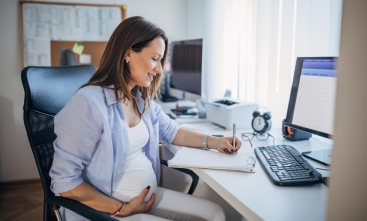 Mulher grávida, sentada, de perfil, trabalhando em um notebook. Ela segura a barriga com um olhar feliz