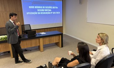 Homem de terno cinza faz uma apresentação a duas mulheres sentadas em um auditório