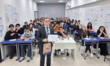 Homem de terno mostrando um certificado em uma sala de aula com diversos adolescentes sentados em carteiras escolares.