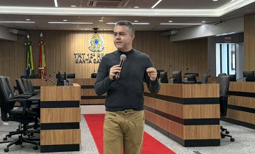 Homem de blusa preta e calça caqui, de pé, fala ao microfone. Ele está em um auditório, tendo ao fundo um brasão do Poder Judiciário iluminado