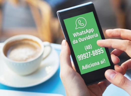 Foto de uma xícara de café expresso ao fundo e na frente uma mão segurando um celular smartphone com o texto: whatsapp da ouvidoria 48 99927-8895, adiciona ai. 