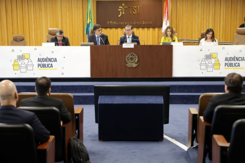 Três homens e duas mulheres compondo uma mesa de evento, em um auditório de paredes amarelas