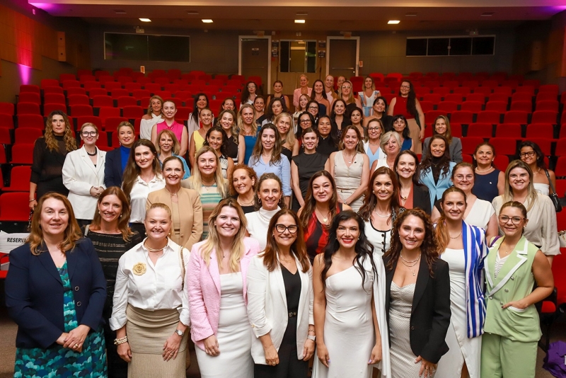 Grupo de cerca de 50 mulheres posam para foto em um amplo auditório, com poltronas vermelhas