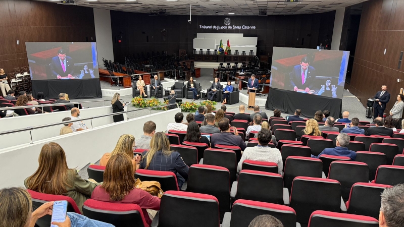 Auditório do TJSC visto do último degrau. Dois telões, um em cada lado. As cadeiras, vermelhas, são separadas por um corredor de entrada