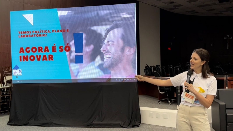 Mulher de cerca de 40 anos falando ao microfone e vestindo uma roupa mais informal. Ela está apontando para uma telão onde se ele: Agora é só inovar