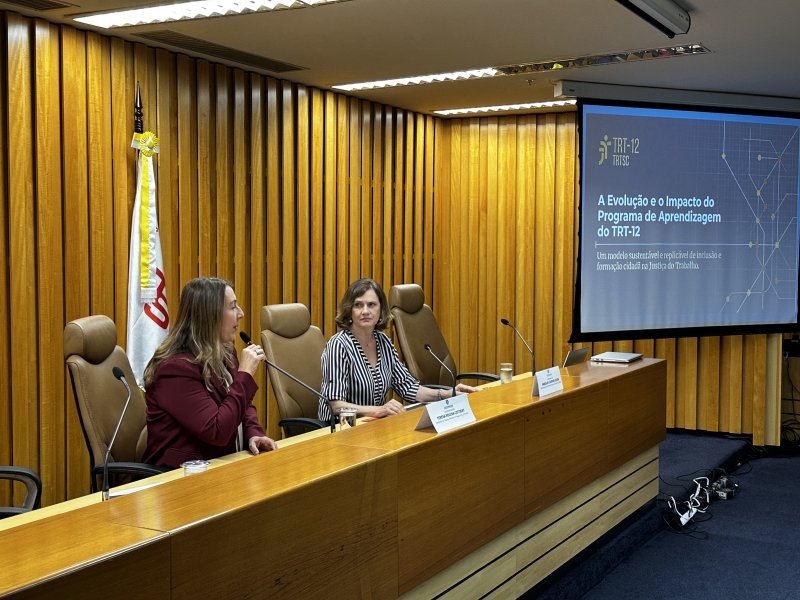 Duas magistradas em uma mesa de honra; uma delas fala ao microfone