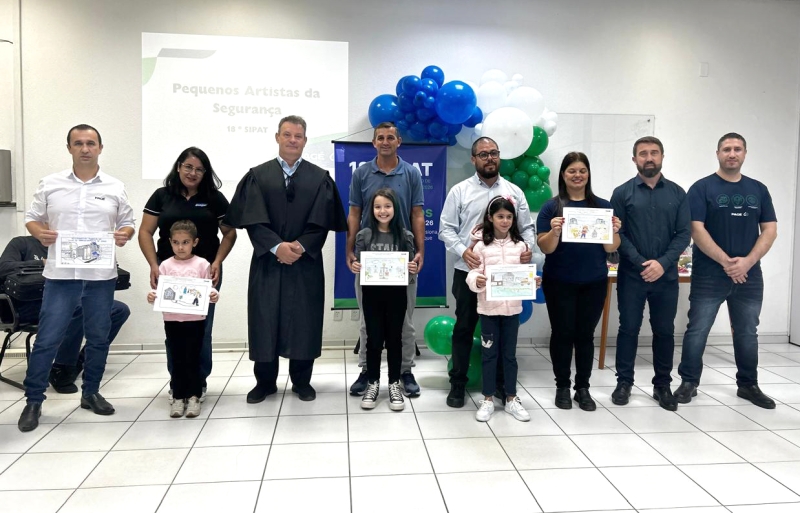 Oito adultos e três crianças posam para foto. Os adultos estão lado a lado, e as crianças, na frente dos pais, exibem desenhos. Ao fundos, balões azuis e um banner.