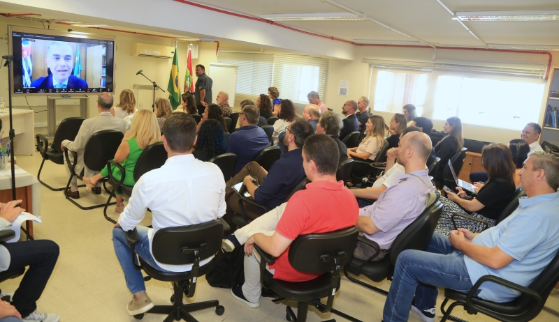 Grupo de 30 pessoas acompanham conferência por vídeo em uma sala de aula