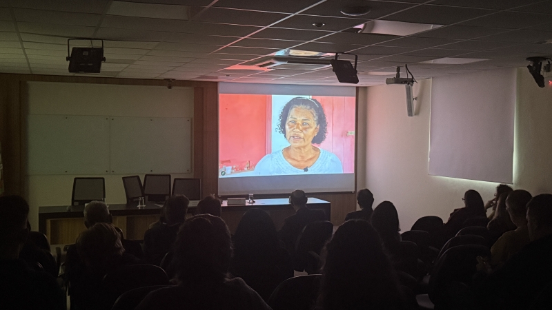 Foto de um auditório escuro, no qual aparece a imagem de uma cena de um filme, em um telão