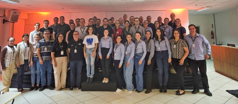 Grupo de cerca de 50 pessoas, todas em pé, posa para a foto em um auditório.