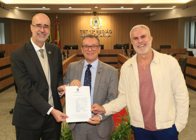 Dois homens de terno e um vestido informalmente posam para foto em uma sala de julgamentos. Todos eles exibem um documento