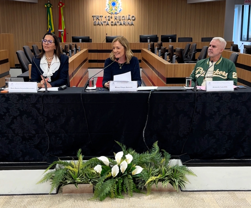 Duas mulheres e um homem compõem a mesa de um evento. Ao fundo, o brasão do Poder Judiciário com o letreiro TRT-SC. Todos estão sorridentes.