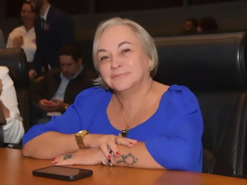 Foto de uma mulher de cabelo grisalho sentada e apoiada em uma mesa, sorrindo para a foto